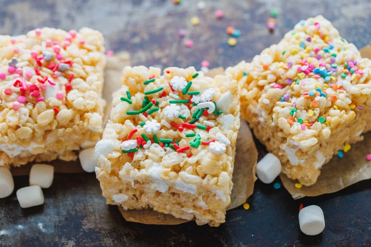 Make thick and gooey Rice Krispie Treats filled with sweet and sticky marshmallows and crispy rice crunch. A family-favorite no-bake dessert! Three Rice Krispie treats decorated with different colored sprinkles next to scattered mini marshmallows.