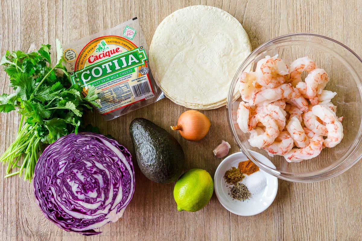 My easy Shrimp Tacos are loaded with shrimp, cabbage slaw, cotija cheese, and the BEST garlic-lime crema sauce. It's the only recipe you need! Shrimp Taco Ingredients with corn tortillas, raw shrimp, seasonings in bowl, avocado, cabbage and cotija cheese