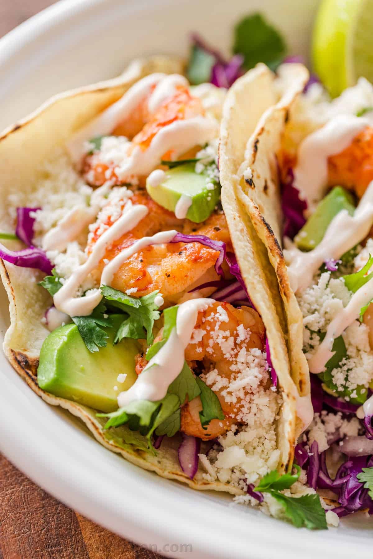 My easy Shrimp Tacos are loaded with shrimp, cabbage slaw, cotija cheese, and the BEST garlic-lime crema sauce. It's the only recipe you need! Side profile of tacos laying on platter loaded with shrimp and drizzled with garlic-lime crema sauce