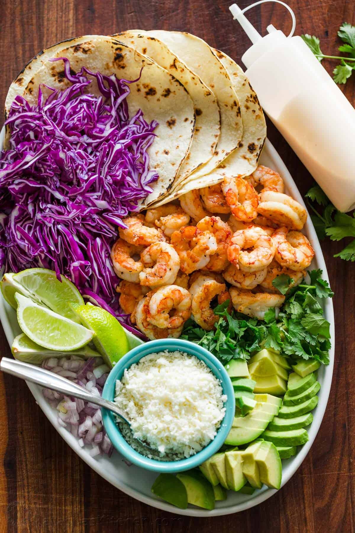 My easy Shrimp Tacos are loaded with shrimp, cabbage slaw, cotija cheese, and the BEST garlic-lime crema sauce. It's the only recipe you need! White serving platter for assembling Mexican seafood dinner cabbage, lime wedges, tortillas, onion, cotija cheese, avocado, and cilantro