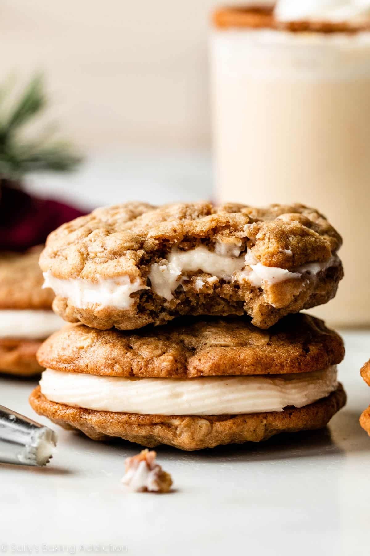 eggnog oatmeal cream pie with bite taken out sitting on top of another whole cookie sandwich with glass of eggnog and other cookies in background.