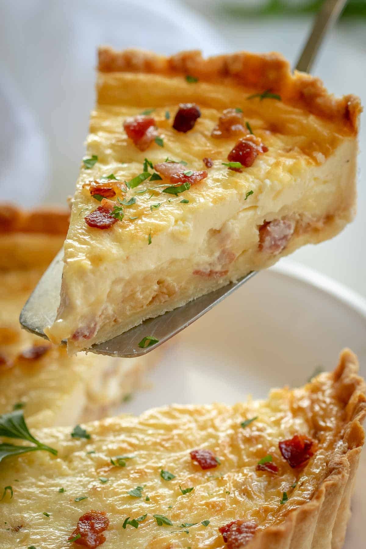 Classic Quiche Lorraine with a savory egg custard, bacon, and cheese filling in flaky pie crust. Quiche Lorraine is the gold standard of quiche recipes! Perfect breakfast and brunch food. slice of quiche Lorraine on a pie server
