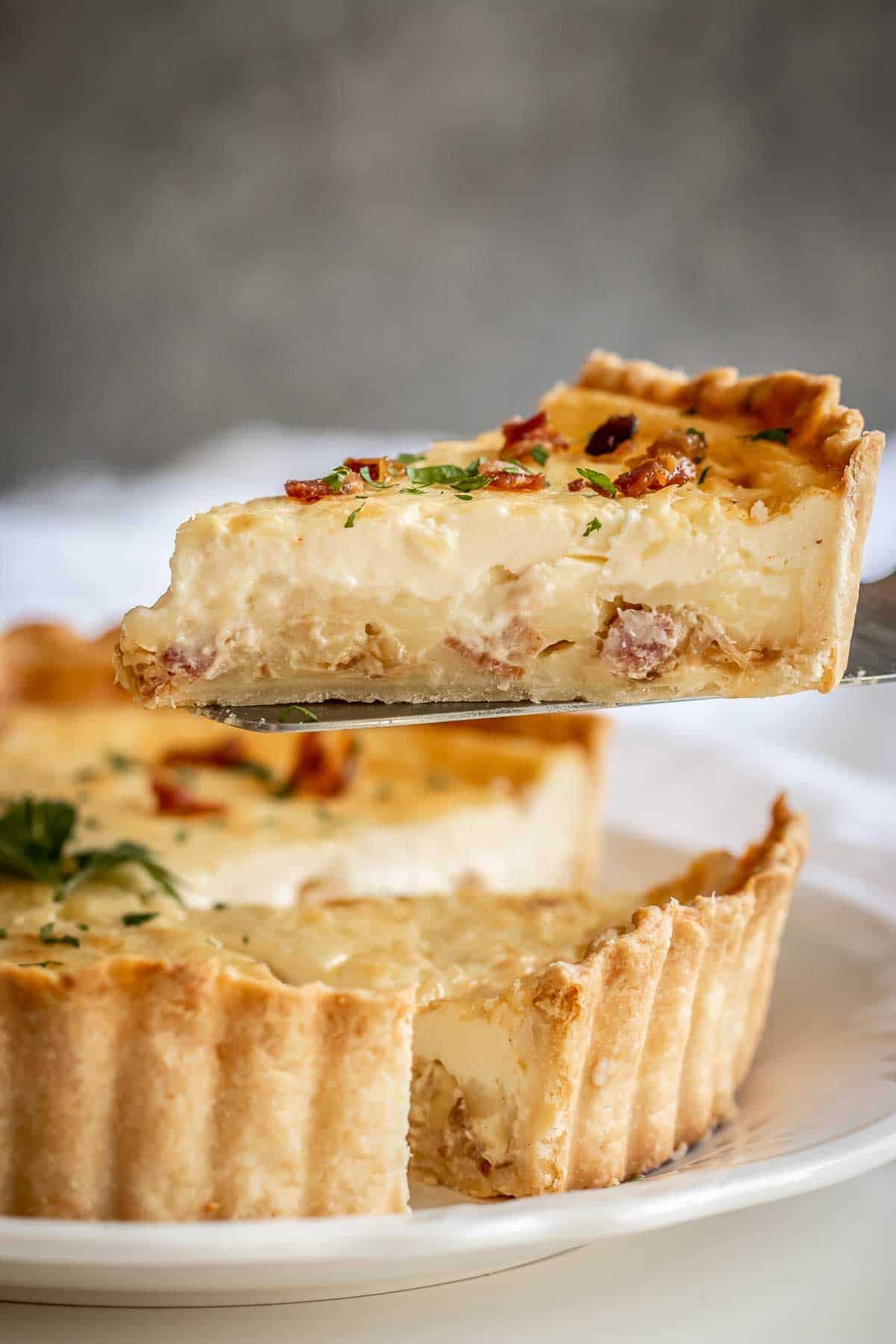 Classic Quiche Lorraine is a delicious breakfast tart that has the best morning foods - eggs, bacon and cheese. A slice of classic quiche Lorraine slice raised above the tart.