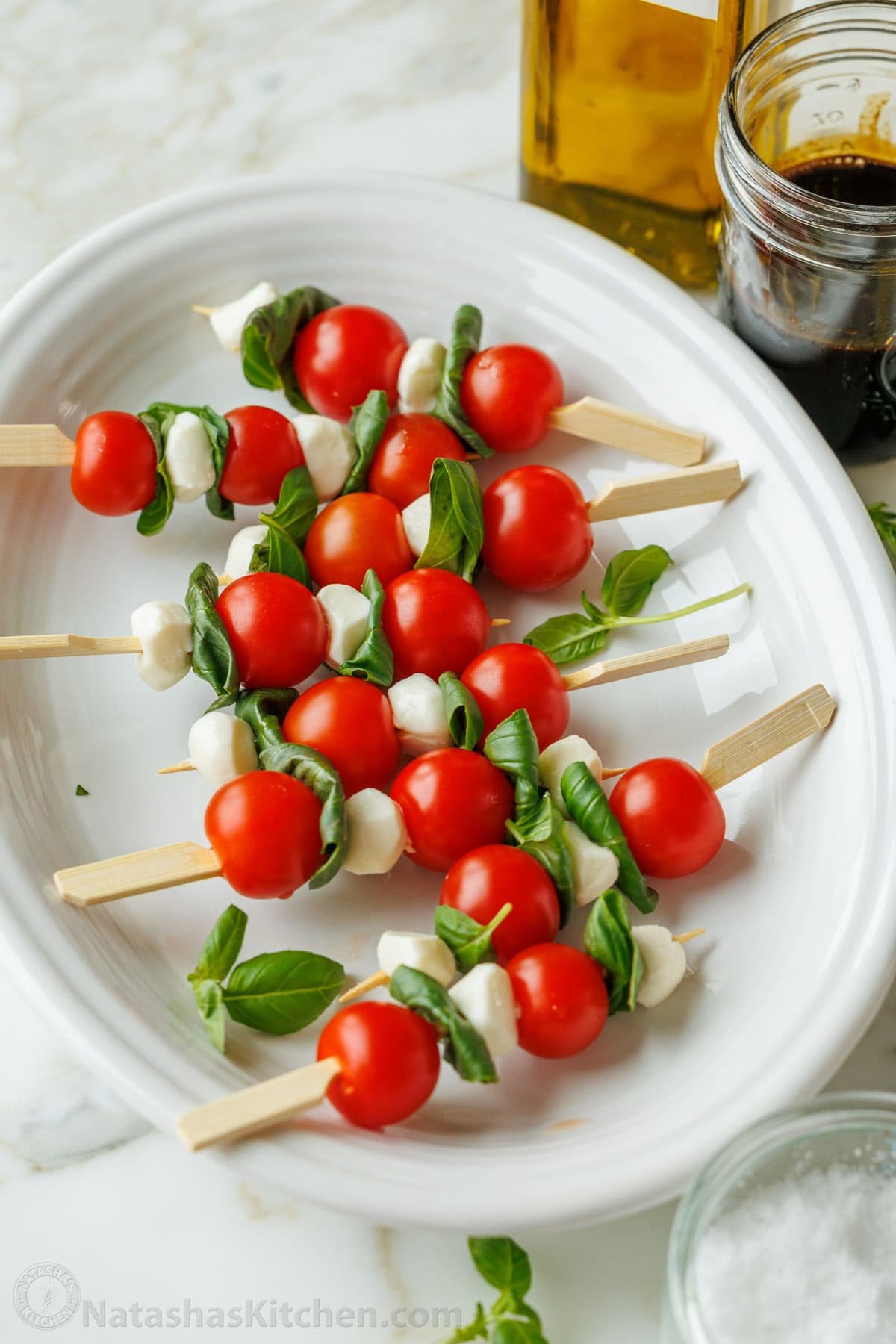 Caprese Skewers is the bite-sized version of the classic Caprese Salad. It takes 10 minutes and 4 ingredients to make these fancy appetizers! Caprese skewers on a white plate with balsamic glaze in a jar