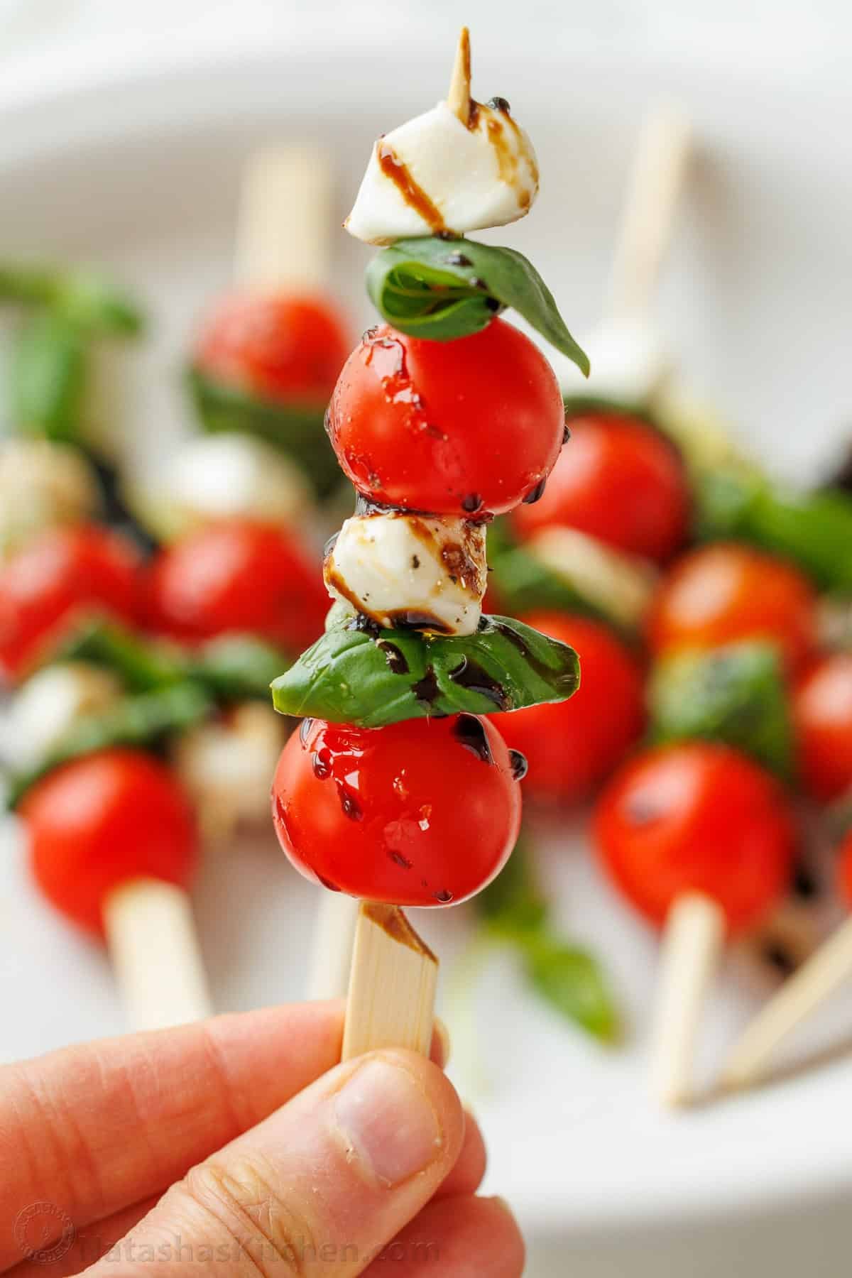 Caprese Skewers is the bite-sized version of the classic Caprese Salad. It takes 10 minutes and 4 ingredients to make these fancy appetizers! a hand holding a caprese skewer appetizer above a tray of skewers
