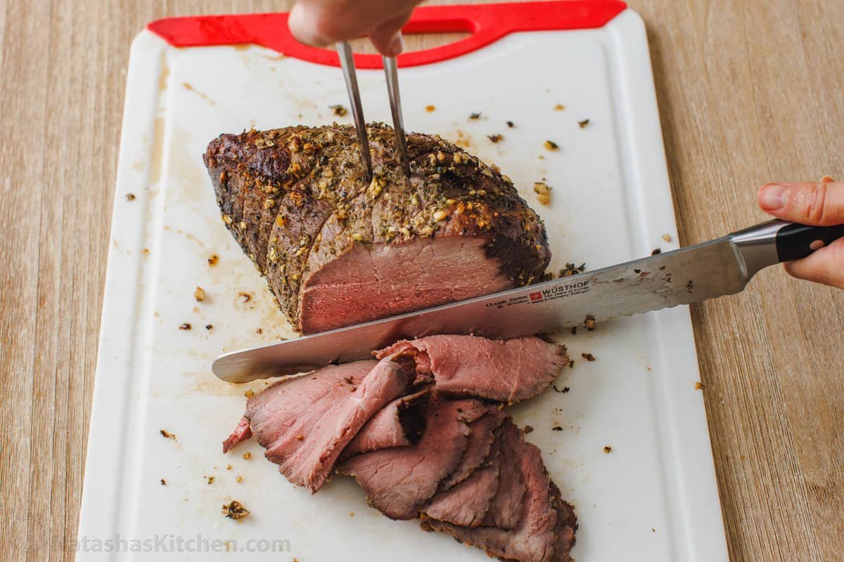 This simple, juicy Roast Beef with savory homemade pan gravy is just as impressive as any holiday roast. Every bite melts in your mouth. A carving fork stuck into roast beef on a cutting board, while a knife cuts off thin slices.