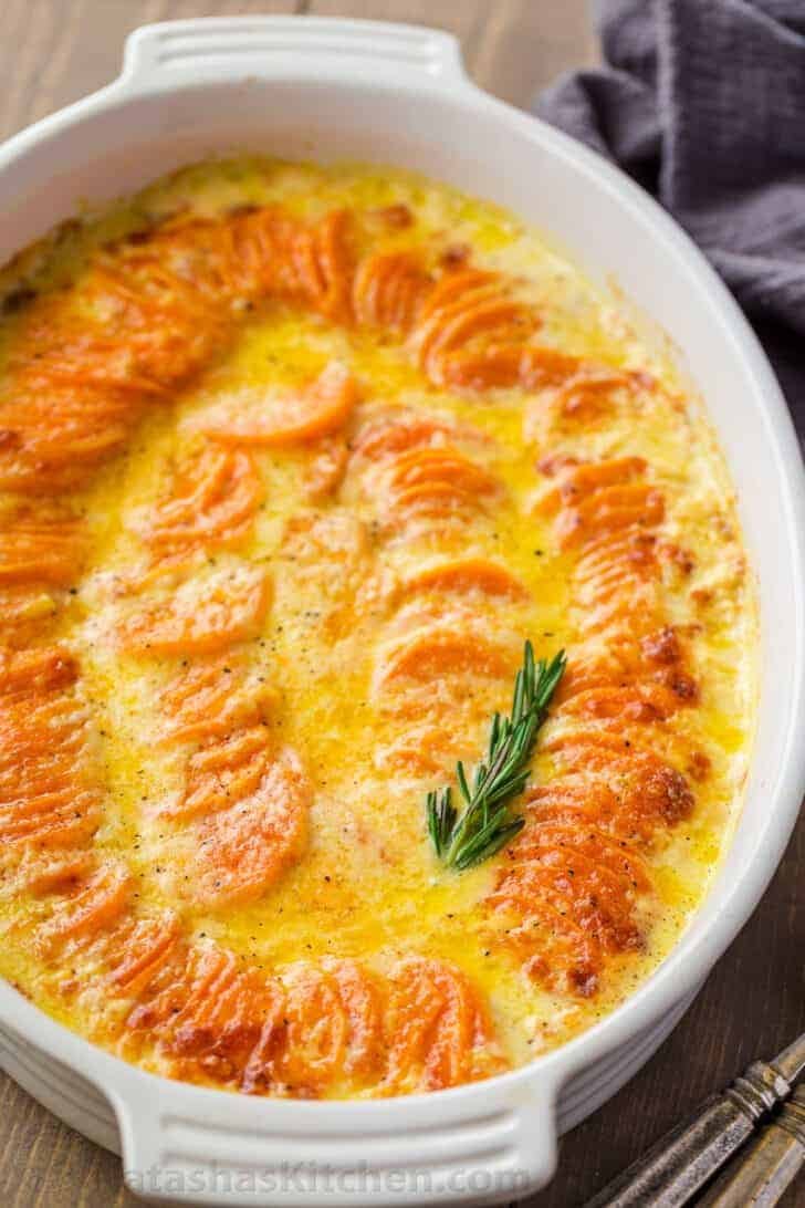 Stacked scalloped sweet potatoes in a casserole dish topped herbs. 