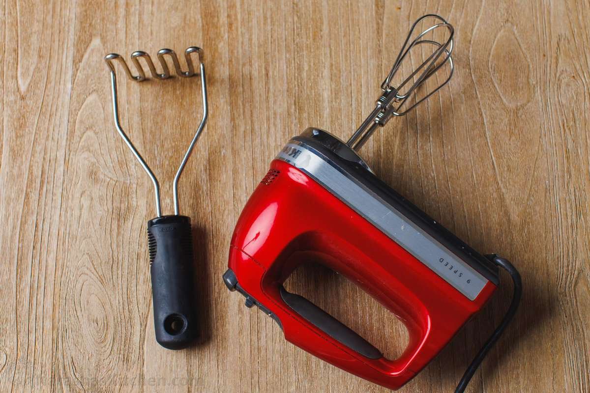 This is a simple sour cream mashed potatoes recipe with loads of buttery flavor. Enjoy homemade mashed potatoes as an easy, versatile side! Overhead view of a potato masher on a countertop next to an electric hand mixer.
