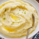 A bowl of sour cream mashed potatoes topped with melted butter and freshly chopped chives, with a spoon.