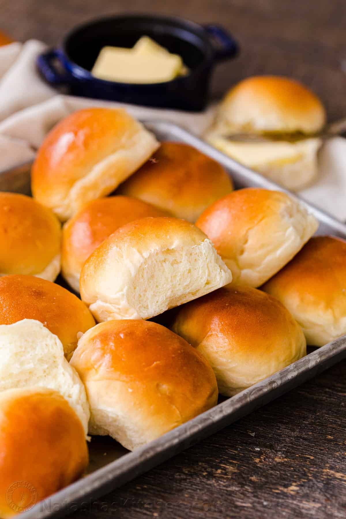 Irresistibly Soft Dinner Rolls are easy to make with just 6 ingredients. There's nothing like fluffy warm homemade dinner rolls. These are holiday worthy! #dinnerrolls #softdinnerrolls #homemadedinnerrolls #easydinnerrolls #howtomakedinnerrolls #natashaskitchen Soft dinner rolls on a tray with butter in the background