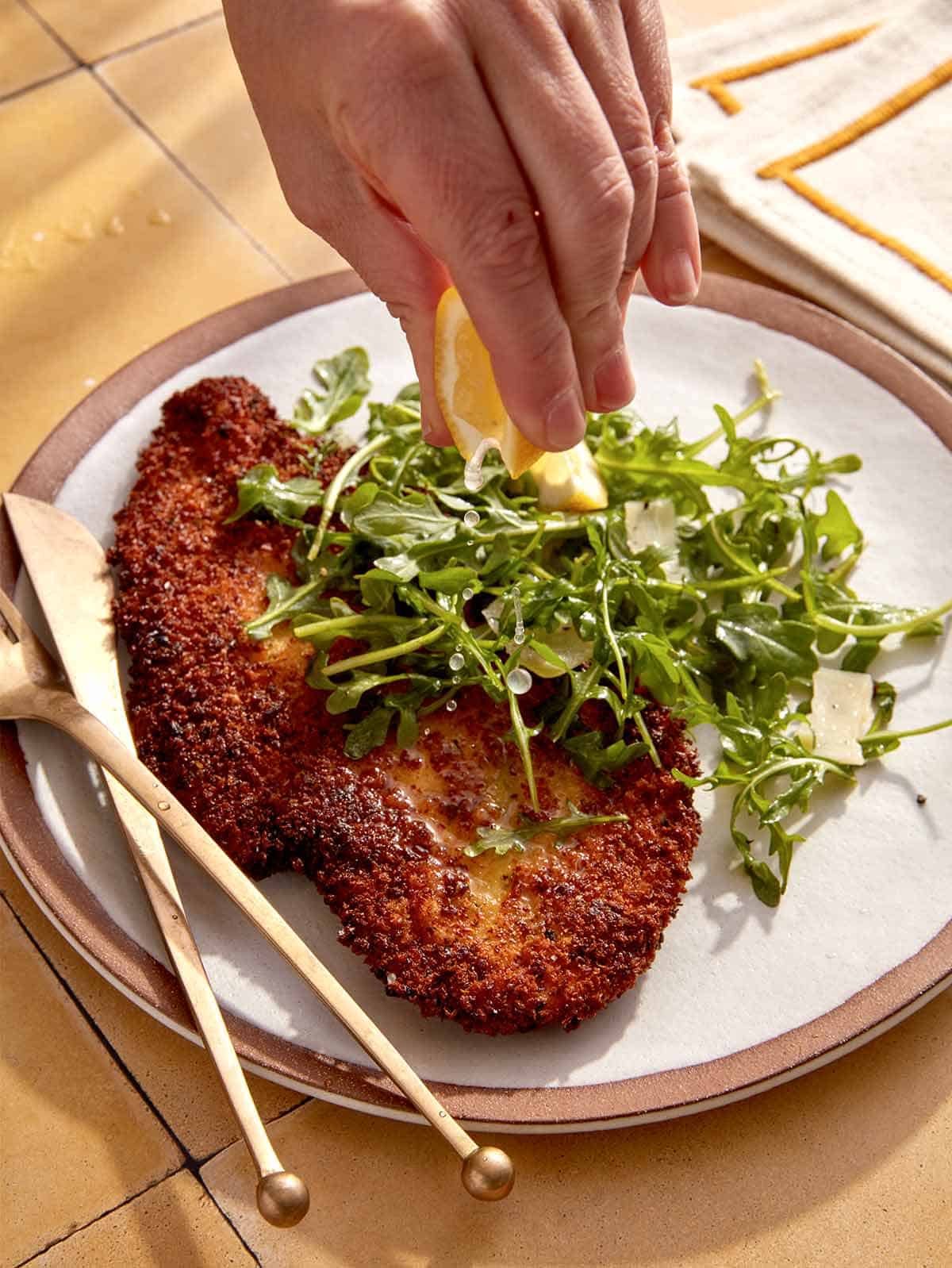 Chicken milanese on a dinner plate with someone squeezing lemon on it.