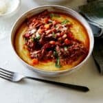 A bowl of braised beef ragu with a fork and a bowl of parmesan on the side.