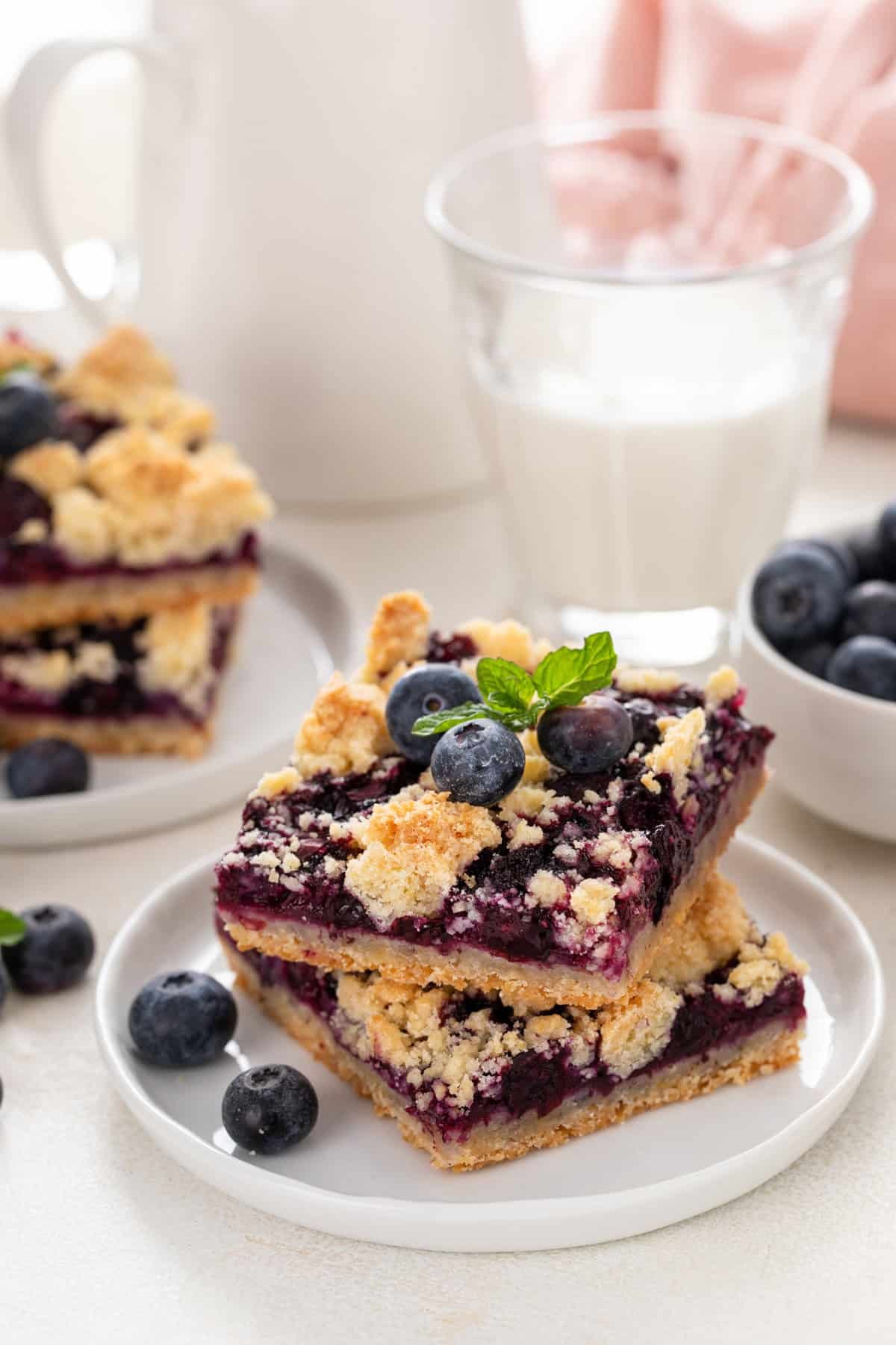 Stacked blueberry crumb bars on white plates in front of a glass of milk.