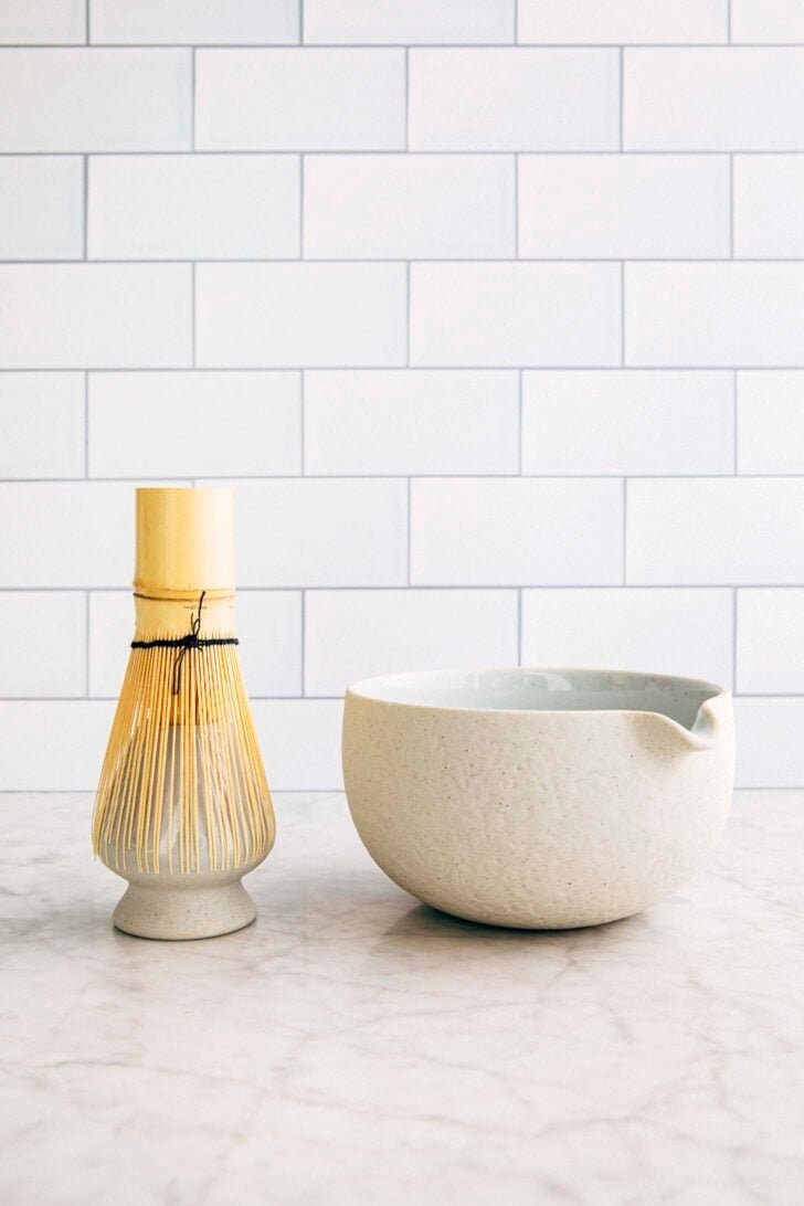 photo of a matcha whisk and a matcha bowl with a spout against a subway tile background