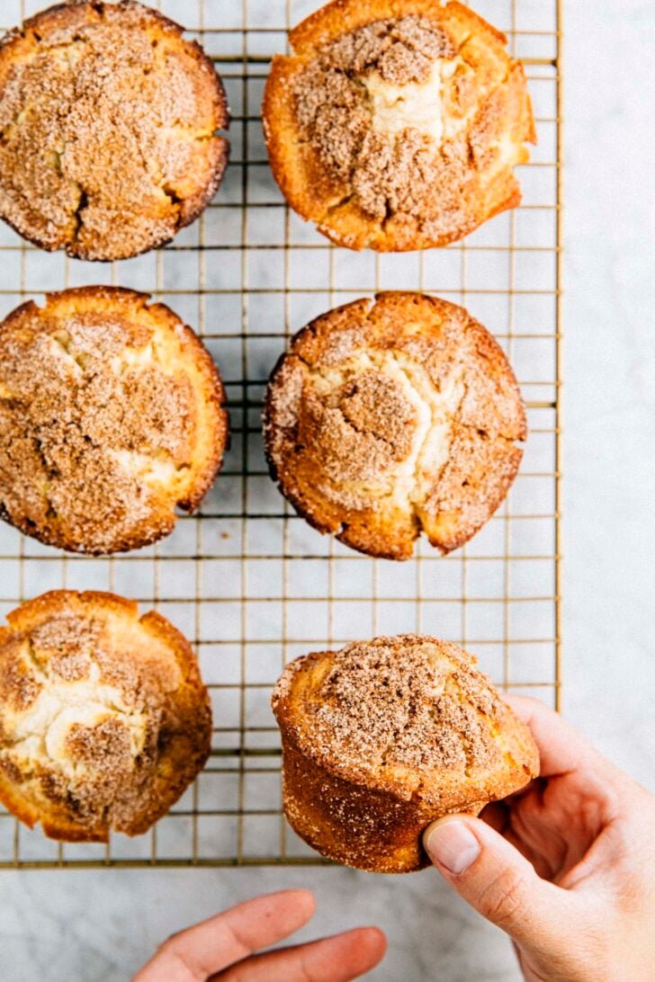 hand picking up a snickerdoodle muffin from a gold wire rack on a marble tabletop