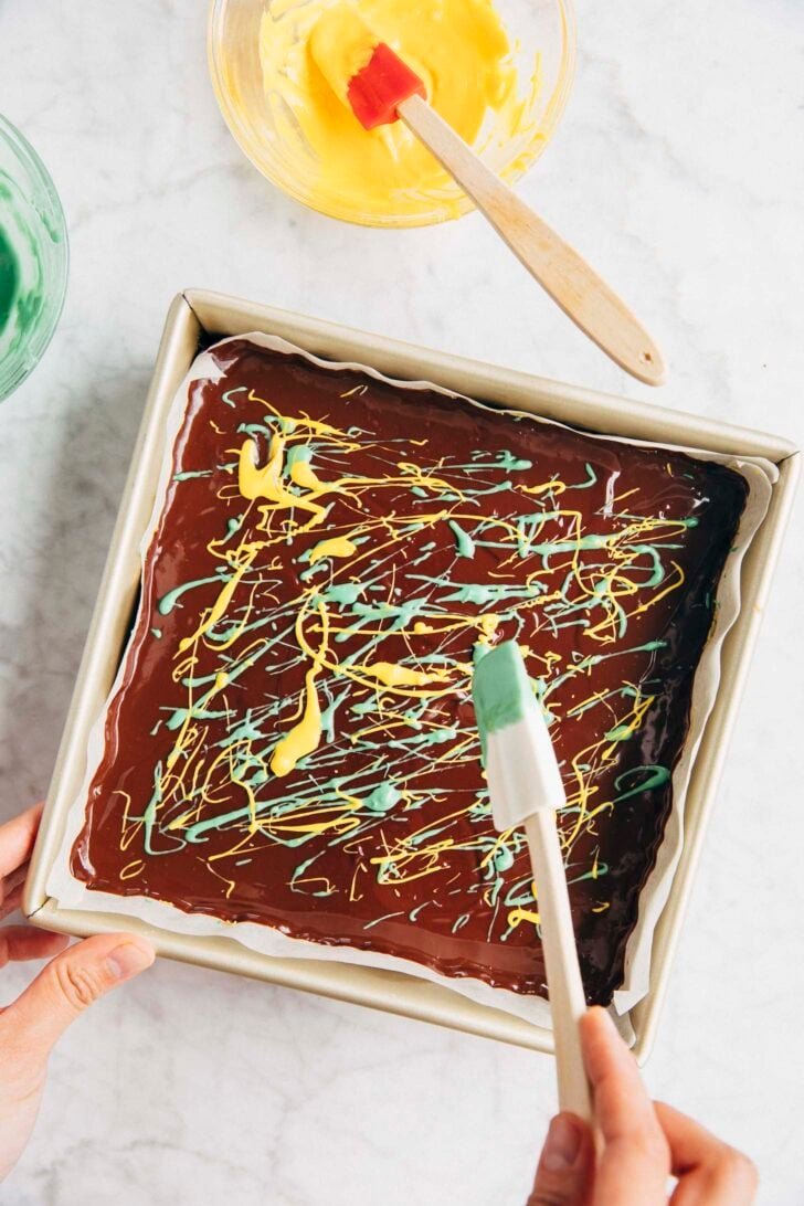 Photo of Michelle from Hummingbird High using a small spatula to drizzle green and yellow candy melts over the ganache layer of the shortbread bars