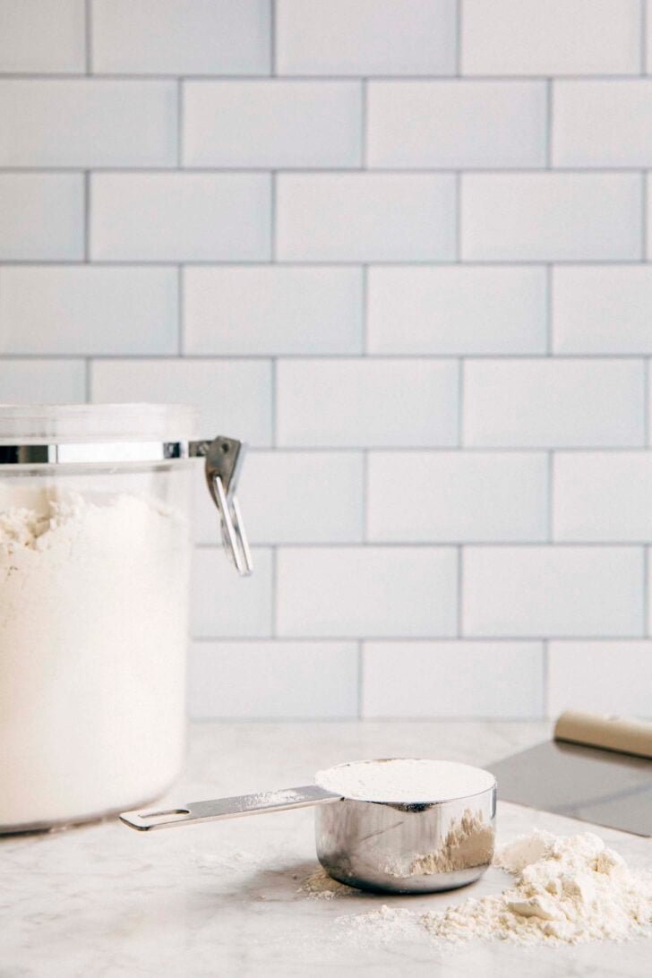 Photo of a measuring cup filled with leveled flour against a white tile backdrop