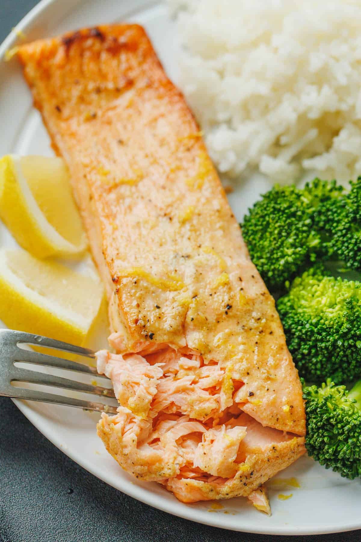 Air Fryer Salmon is one of the best ways to cook salmon in 10 minutes! The air fryer creates a crisp edge with a flaky and juicy center. A fork flakes the end of an air fryer salmon fillet on a white plate served with broccoli and rice.