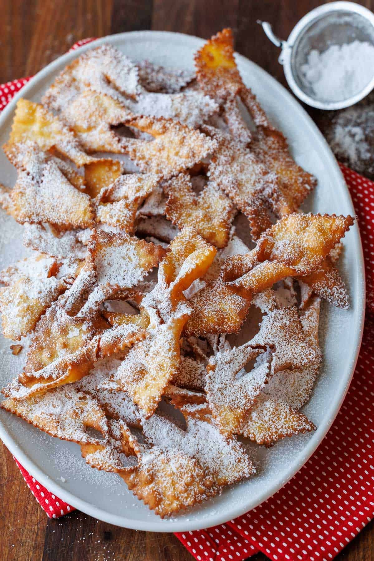 Traditional Angel Wing Cookies (Polish Chrusciki) make a beautiful holiday cookie. These ribbon cookies are dusted with sugar & irresistible. Angel Wing Cookies on a white platter with red and white napkins