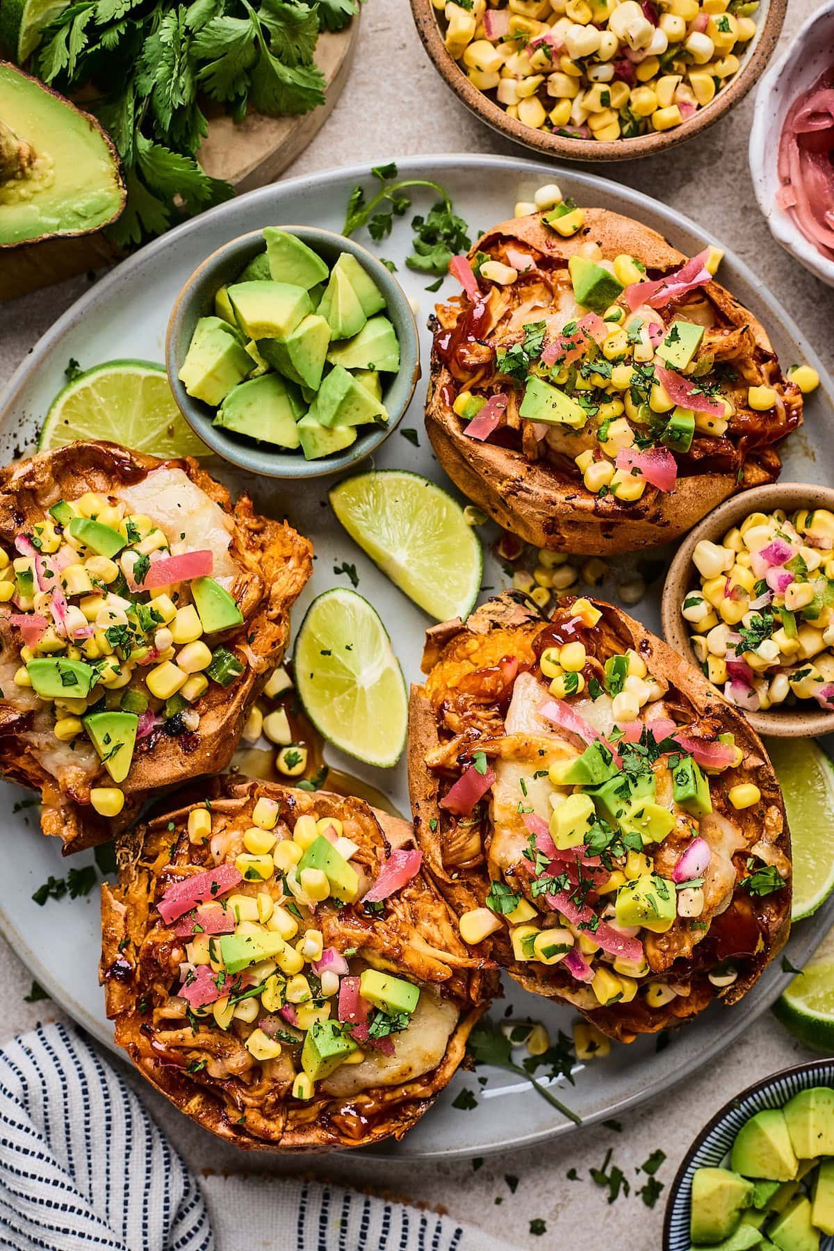 BBQ Chicken Stuffed Sweet Potatoes with corn salsa, pickled red onions, avocado, and lime wedges on plate.