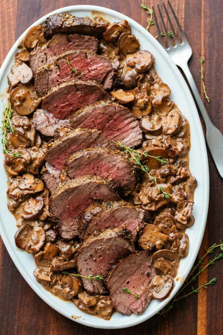 Beef Tenderloin with Mushroom Sauce is a holiday-worthy and impressive main course. Learn how to prep, tie, and cook a beef tenderloin roast in the oven. Sliced beef tenderloin with mushrooms on serving platter with thyme