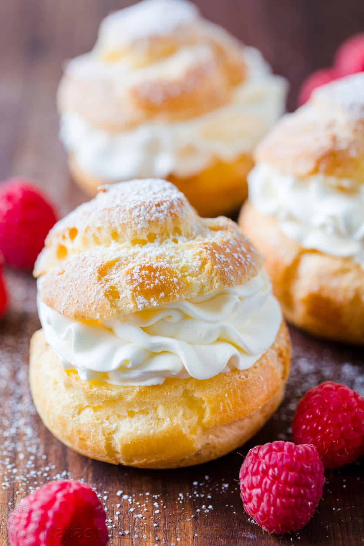 Cream Puffs are a classic French dessert filled with sweet cream and dusted with powdered sugar. Learn how to make easy bakery quality cream puffs. #creampuffs #creampuffrecipe #chouxpastry #chouxpaste #dessert #pastry #Frenchpastry #French #natashaskitchen Cream Puffs filled with cream on cutting board dusted with powdered sugar and garnished with raspberries