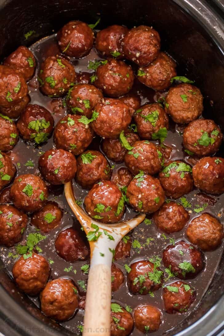 Grape Jelly Meatballs are just frozen meatballs tossed together with grape jelly, bbq sauce, and garlic chili sauce in a crockpot and cooked until meatballs are tender so flavorful! A crockpot full of grape jelly meatballs topped with chopped greens and a ladle.