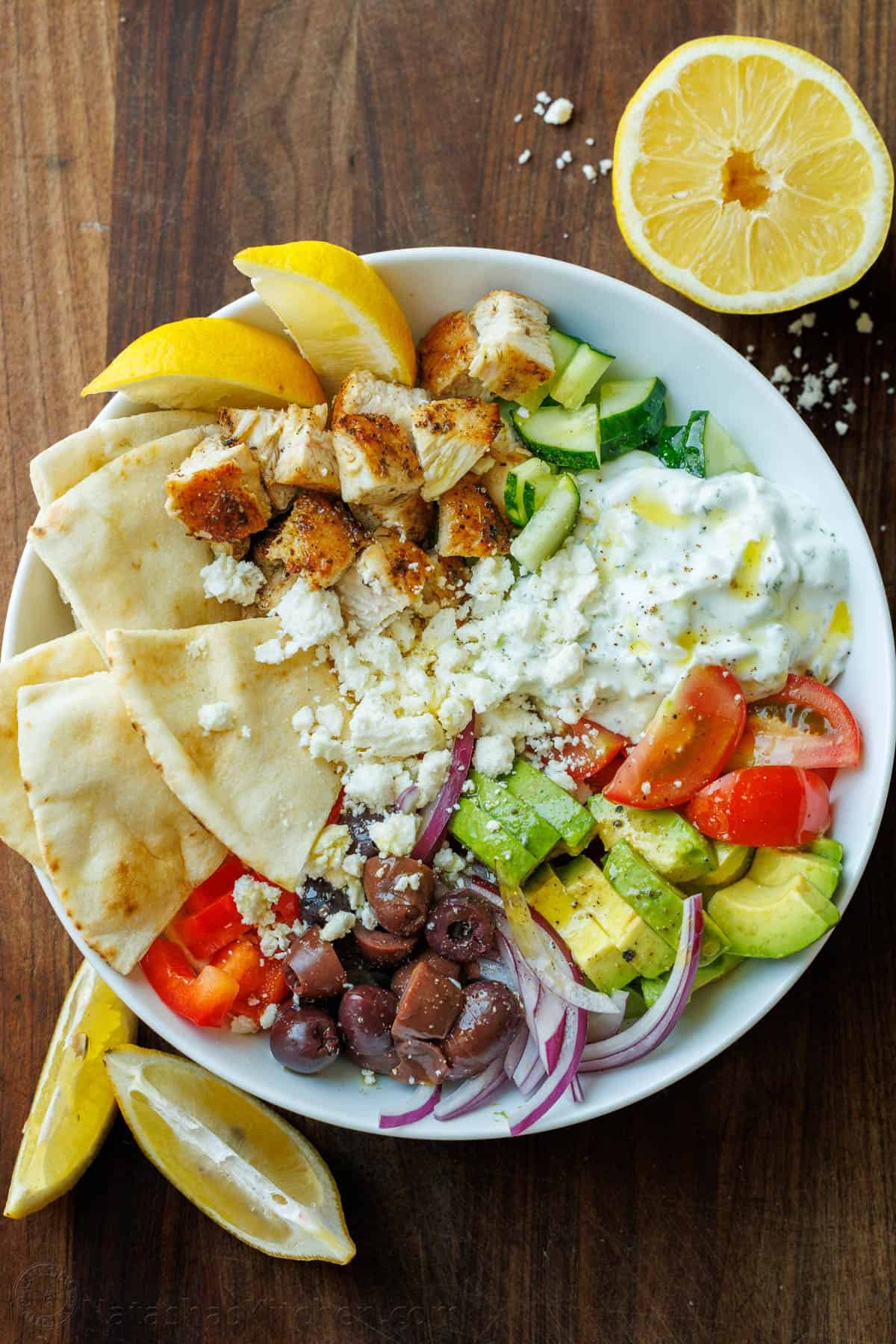 Greek Chicken Bowls combine the classic elements of a Greek Salad—cucumbers, tomatoes, olives, and feta—elevated with juicy grilled chicken and a refreshing dollop of tzatziki. Chopped Greek Chicken Bowl ingredients in a white bowl with lemon wedges and pita bread