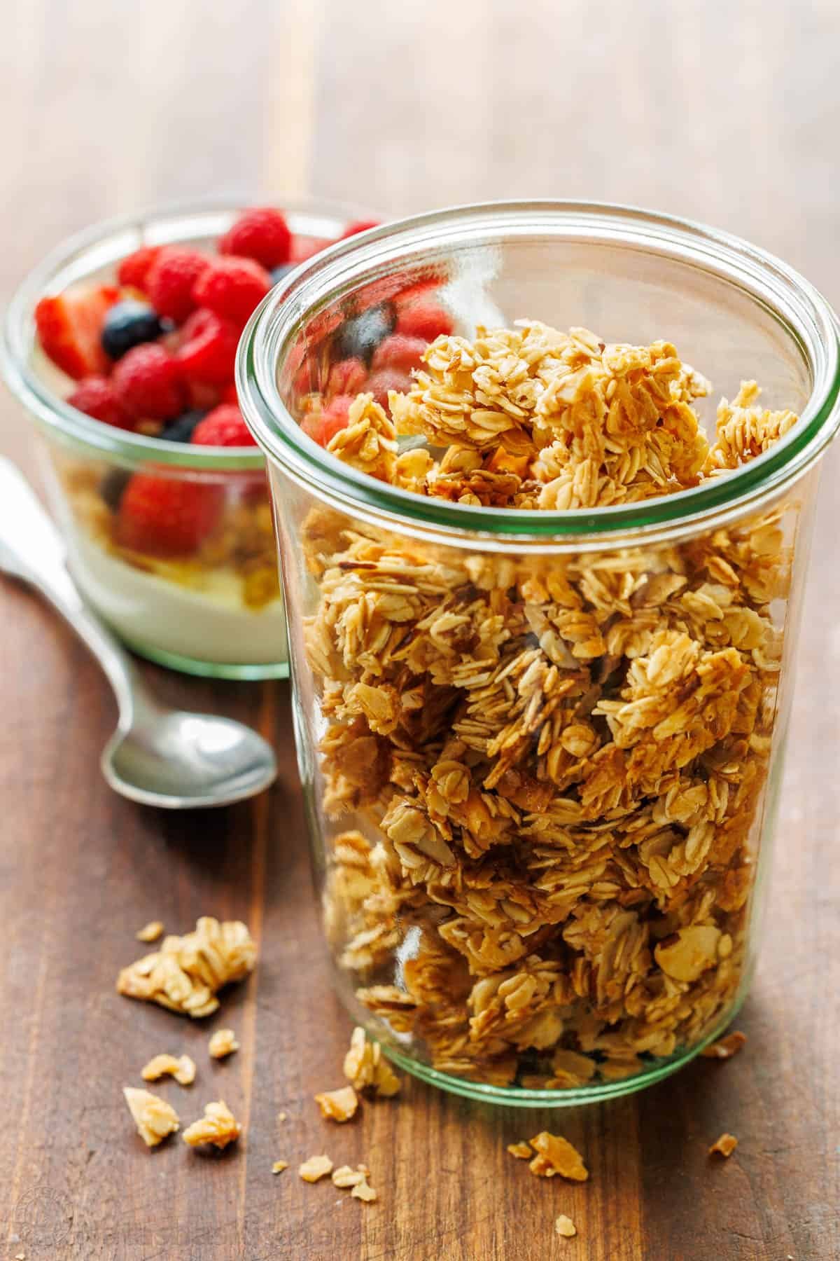 This Homemade Granola Recipe is one of my family's go-to healthy breakfast recipes because it's quick to make and ready to eat all week long. healthy Homemade granola in a glass jar near a yogurt parfait with raspberries and a spoon