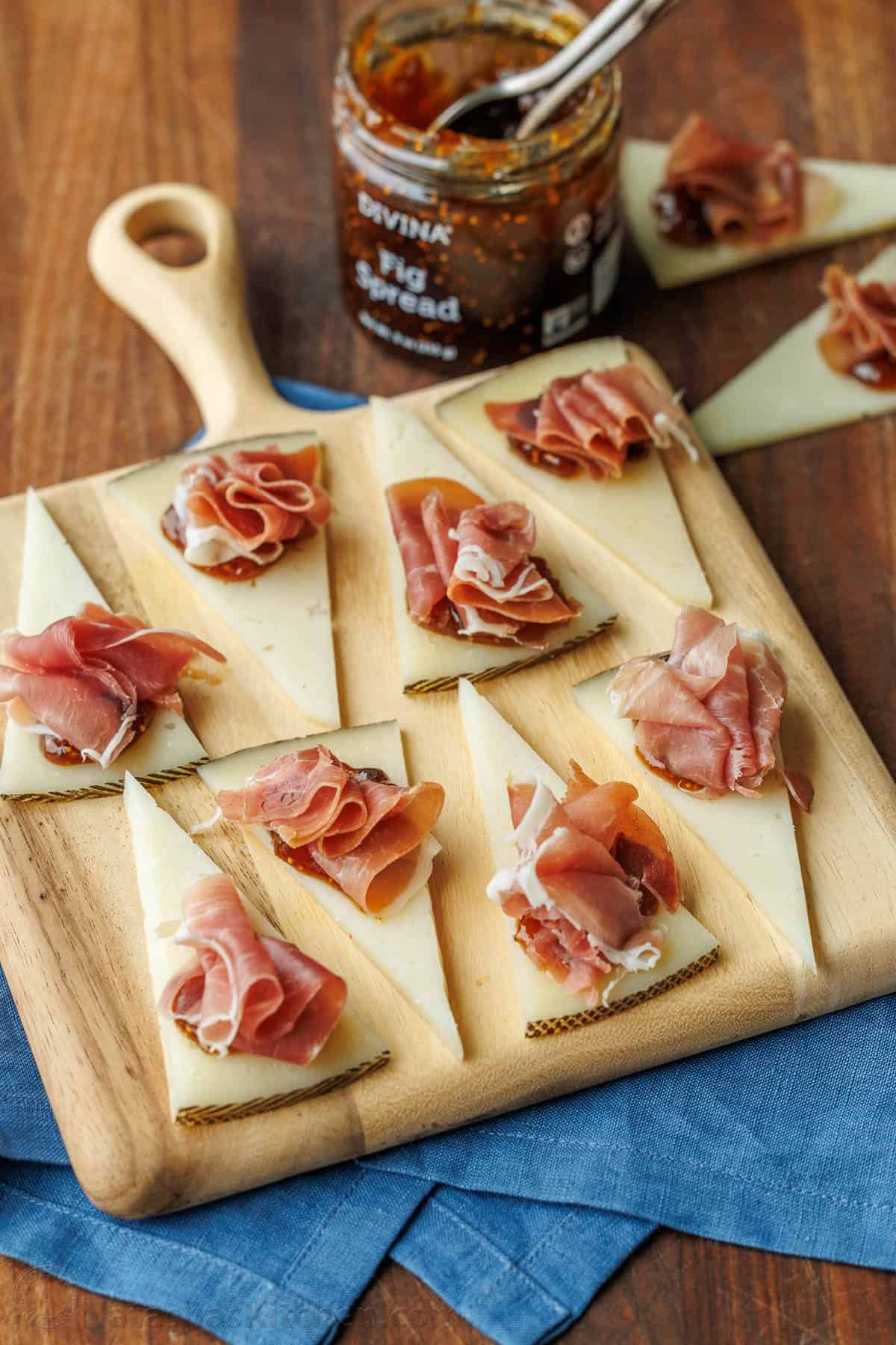 Easy manchego cheese bites with fig preserves and prosciutto take less than 5 minutes to make, but they look elegant and taste divine! Manchego Cheese Bites on a cutting board with fig spread in the background