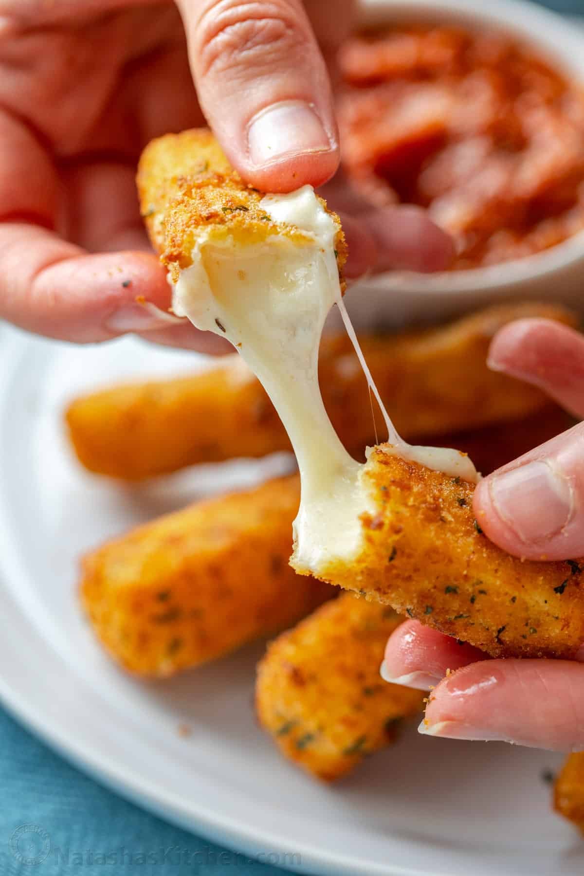 Homemade mozzarella sticks are irresistibly crispy on the outside and gooey and melty in the middle. The ultimate snack or party appetizer! Two hands pulling apart a mozzarella stick with a plate of cheese sticks in the background.