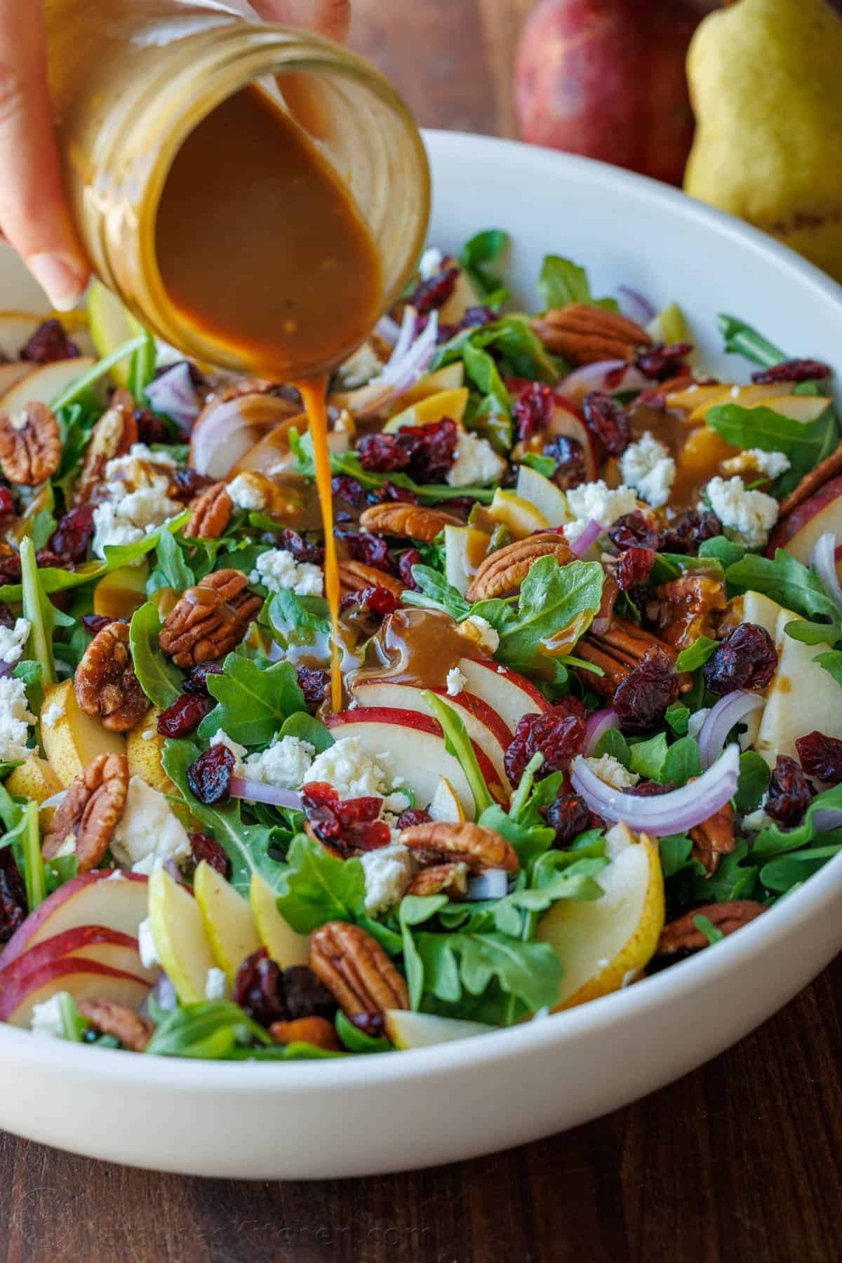 This Autumn Pear Salad is stunning with sweet pears, fresh arugula, tangy Craisins, pecans, goat cheese, and a homemade Balsamic Vinaigrette. Pear Salad being dressed with Balsamic Vinaigrette in a white bowl, loaded with a variety of pears, red onion, goat cheese, cranberries, and toasted pecans