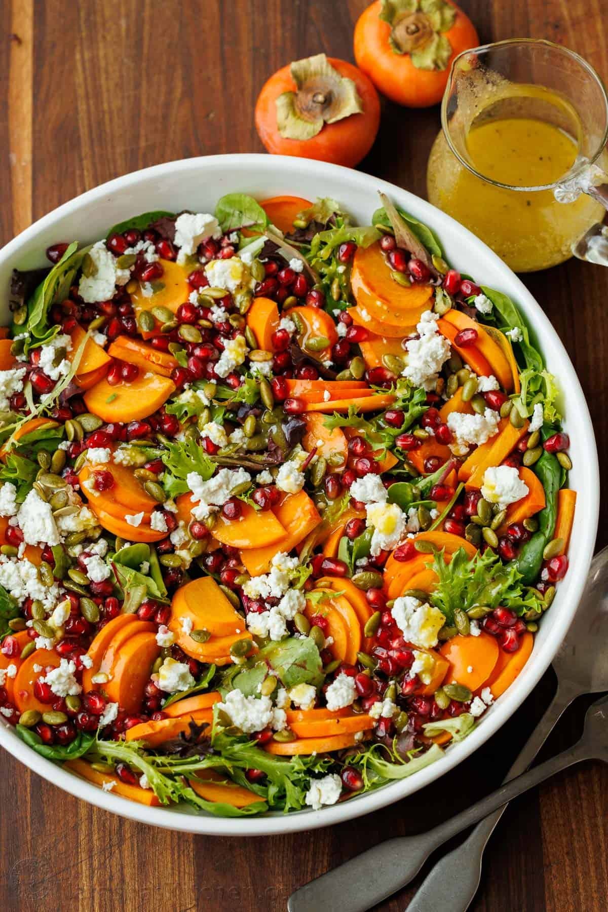 My persimmon Salad recipe features fresh fruit, crunchy toasted pepitas, tangy goat cheese, and a maple vinaigrette. Festive for any holiday! persimmon salad with maple vinaigrette on a wooden table with extra dressing and fruit