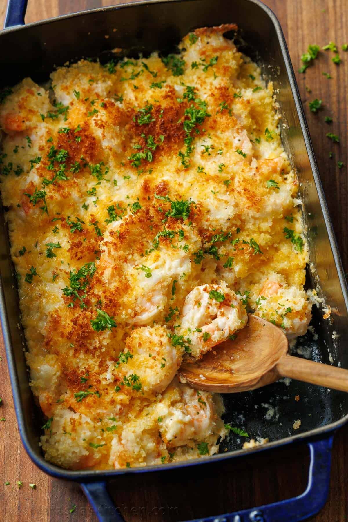 My Shrimp casserole recipe is an easy 30-minute dish for holidays or weeknights alike! Cheesy shrimp filling topped with crunchy breadcrumbs. Shrimp Casserole served out of a dark blue casserole dish with a wooden spoon