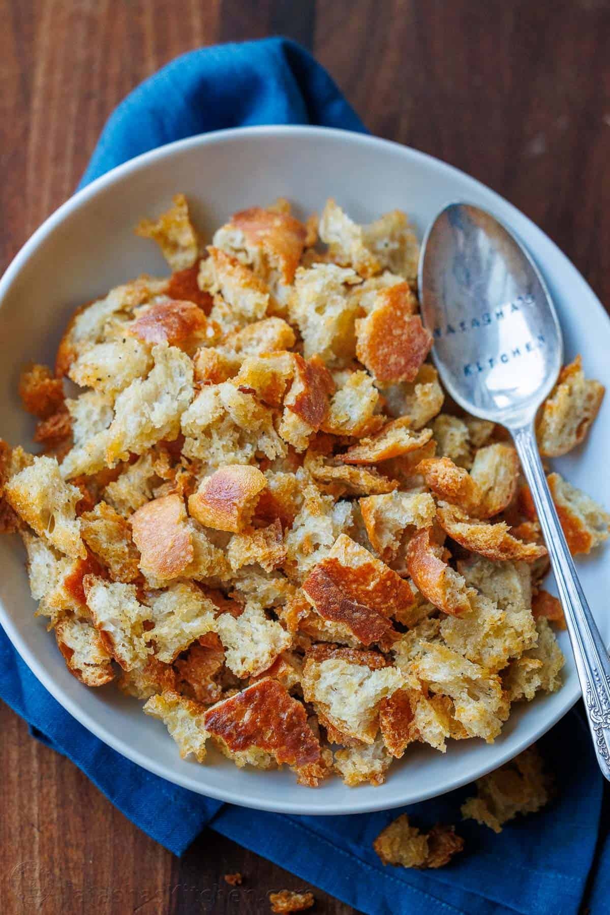 Making Sourdough Croutons is a great way to bring new life to leftover sourdough bread. Use them to bring a delicious crunch to your salads and soups. Sourdough Croutons in a bowl with a metal serving spoon stamped with Natasha's Kitchen