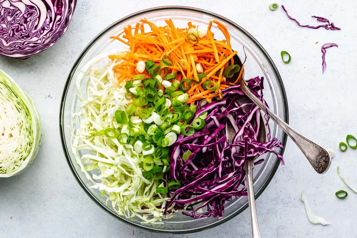 shredded cabbage, carrots, and green onion in mixing bowl with spoons to make coleslaw.
