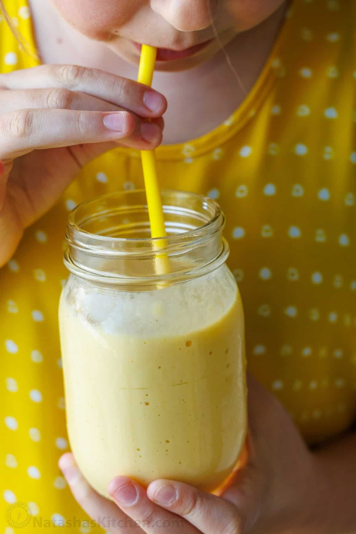 Easy Tropical Mango Smoothie recipe uses just 6 ingredients, like mango, lime, banana, and Greek yogurt, and takes under 5 minutes to make! Small girl drinking a Mango smoothie in a mason jar with a yellow jar