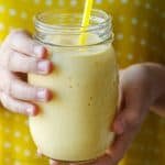 kid's hands holding Mango Smoothie recipe in a mason jar with yellow straw and yellow background