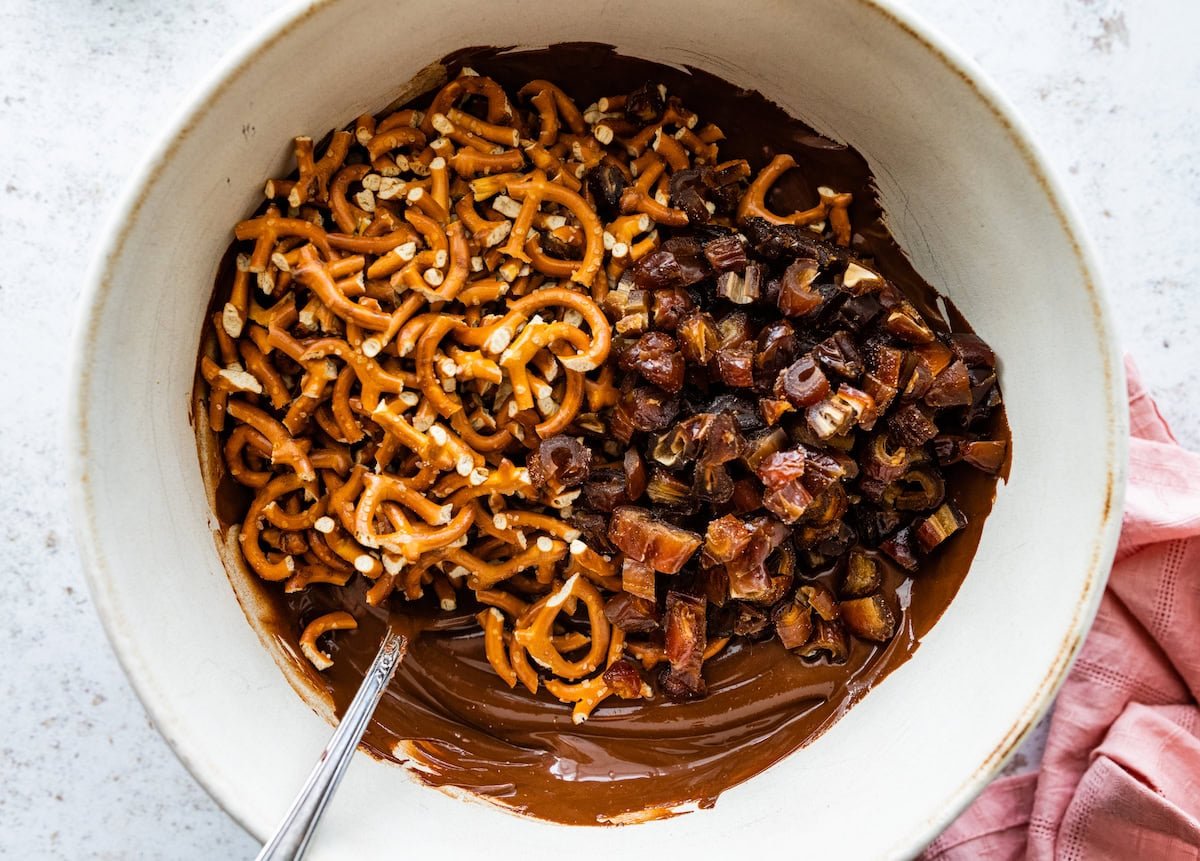 melted chocolate, peanut butter, pretzels, and chopped dates in bowl with spoon.