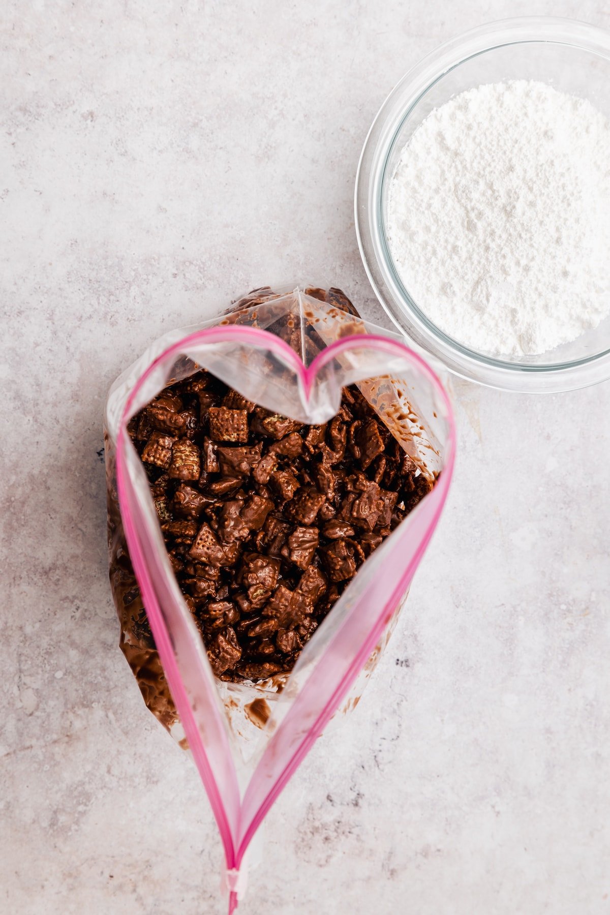 Homemade Puppy Chow is easy to make and SO addicting. This recipe for chocolate peanut butter-covered Chex cereal is a perfect party snack! Overhead view of chocolate and peanut butter-coated Chex cereal in a large ziploc bag, next to a small bowl of powdered sugar.