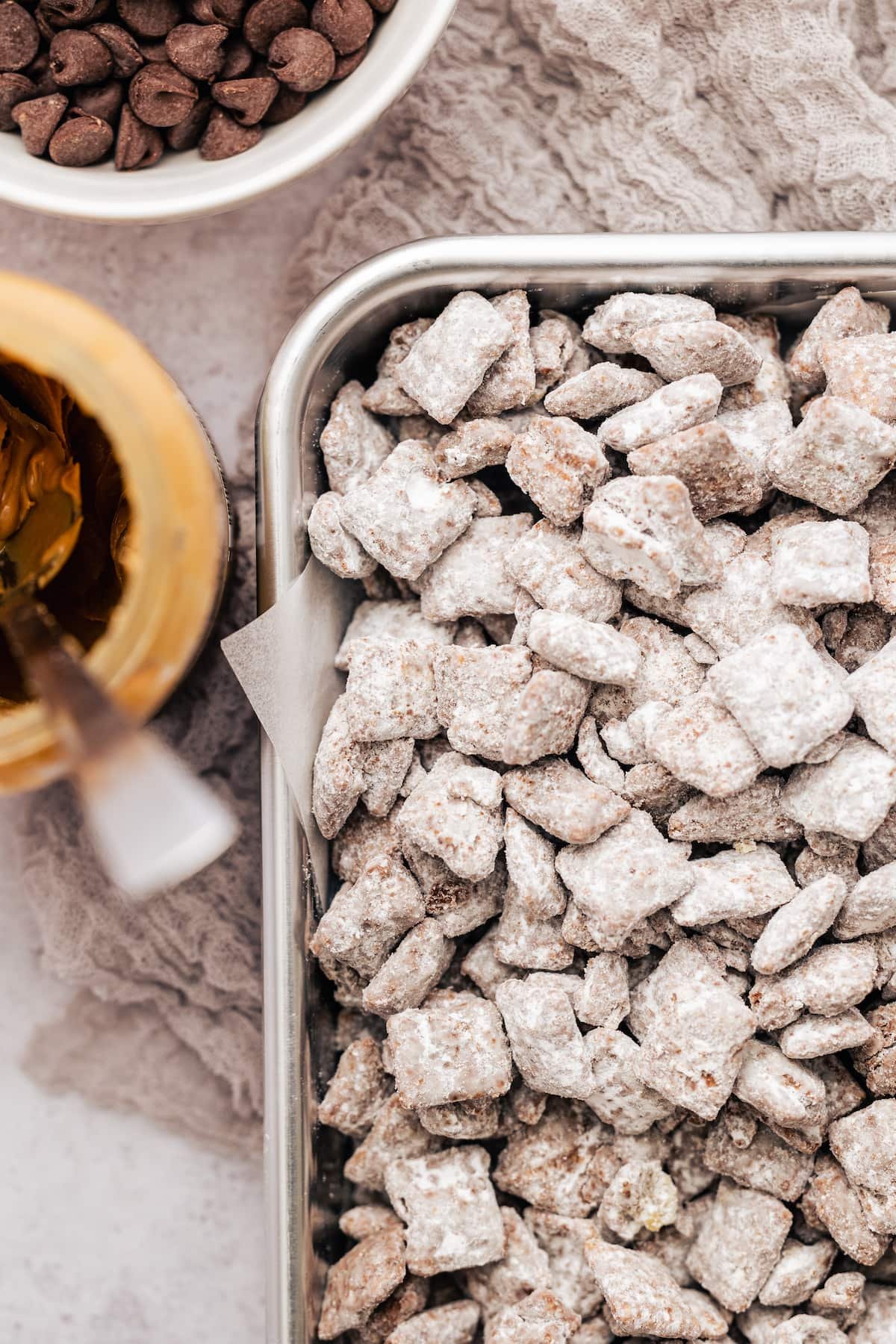 Homemade Puppy Chow is easy to make and SO addicting. This recipe for chocolate peanut butter-covered Chex cereal is a perfect party snack! Overhead view of the corner of a metal pan filled with puppy chow next to a jar of peanut butter and a bowl of chocolate chips.
