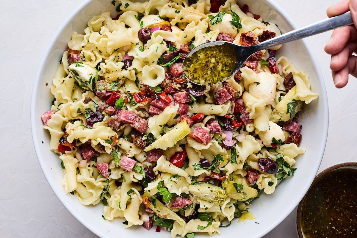 Italian dressing being spooned over Italian pasta salad in serving bowl. 
