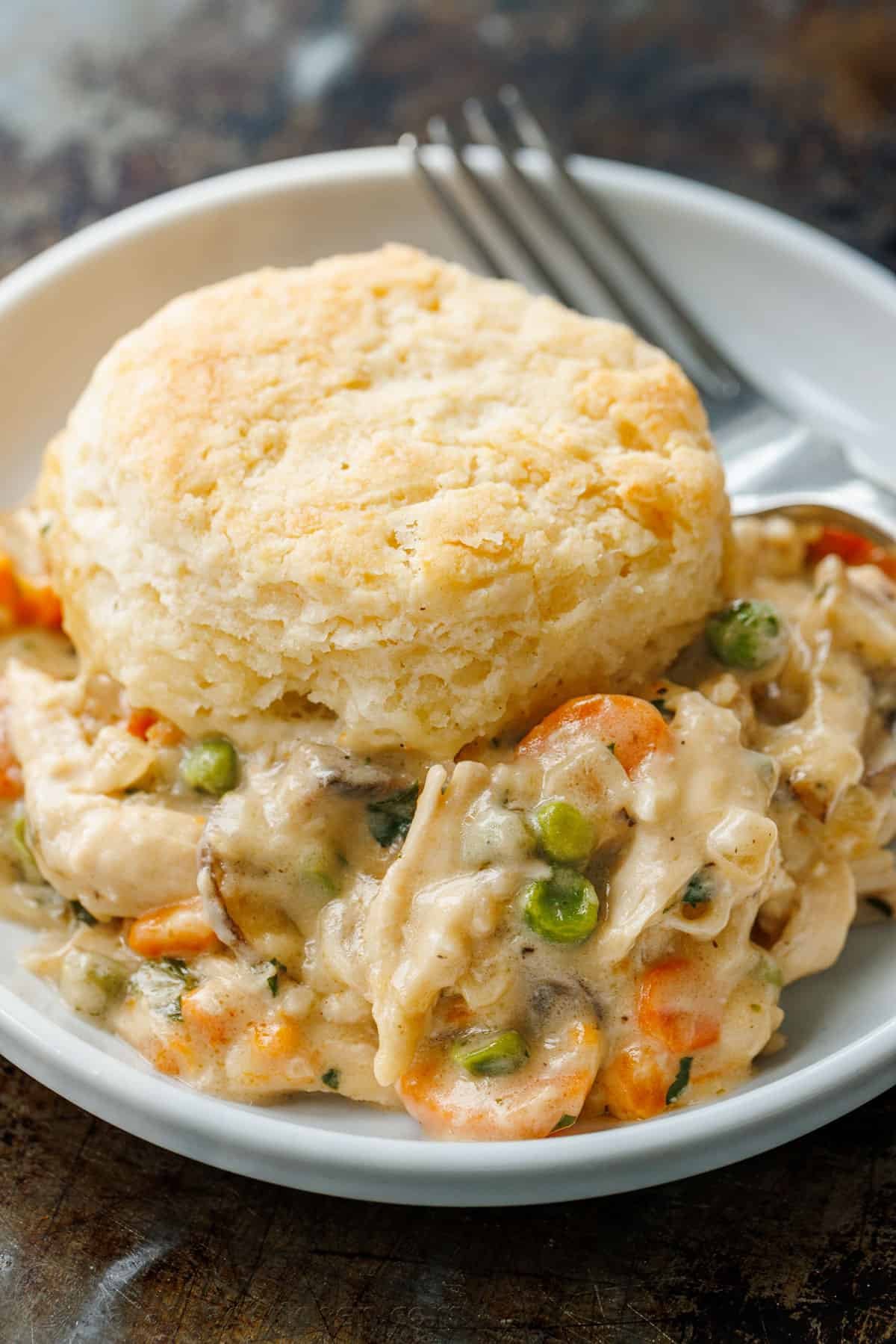 Chicken Pot Pie Casserole has all the cozy flavor of a creamy chicken pot pie, topped with flaky biscuits instead of baked inside a pie crust. A serving of chicken pot pie casserole topped with a biscuit in a bowl, next to a fork.