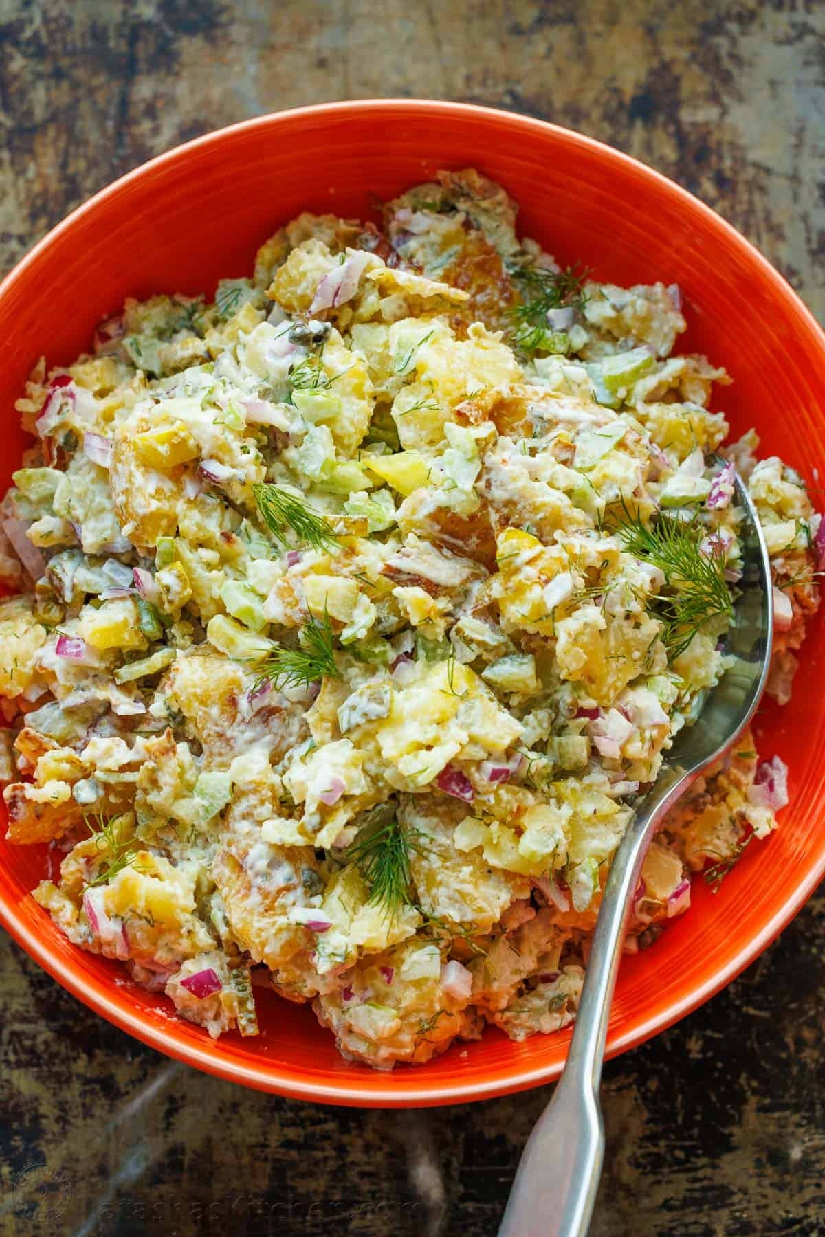Smashed Potato Salad: A crispy twist on potato salad, with crisp vegetables and fresh herbs, combined with an irresistible two-ingredient dressing. This light and satisfying dish will win you over with every bite. Smashed Potato Salad in a red serving bowl with a serving spoon, garnished with fresh dill.