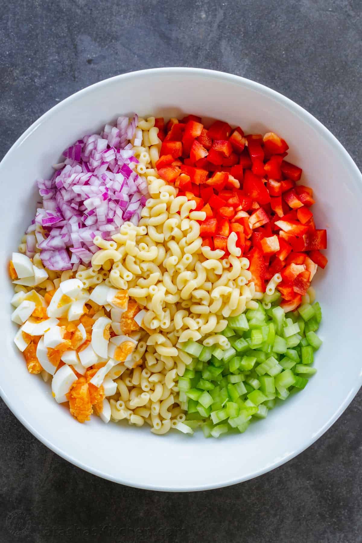 Macaroni Salad is perfect for a summer menu. Paired with BBQ meats, hot dogs, and hamburgers, it's the perfect creamy, refreshing side dish. Ingredients prepped ahead for salad with pasta, bell pepper, celery, egg and onion in a bowl