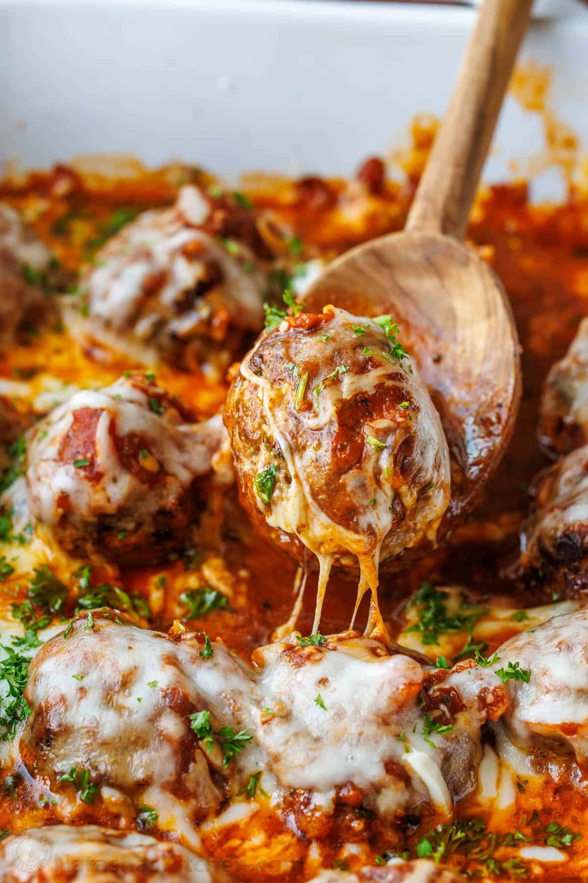 Meatballs Parmesan combines the best Italian dishes into one easy casserole. Homemade saucy Parmesan Meatballs topped with gooey cheese. Serving a homemade meatball from a dish with stringy cheese and parsley garnish