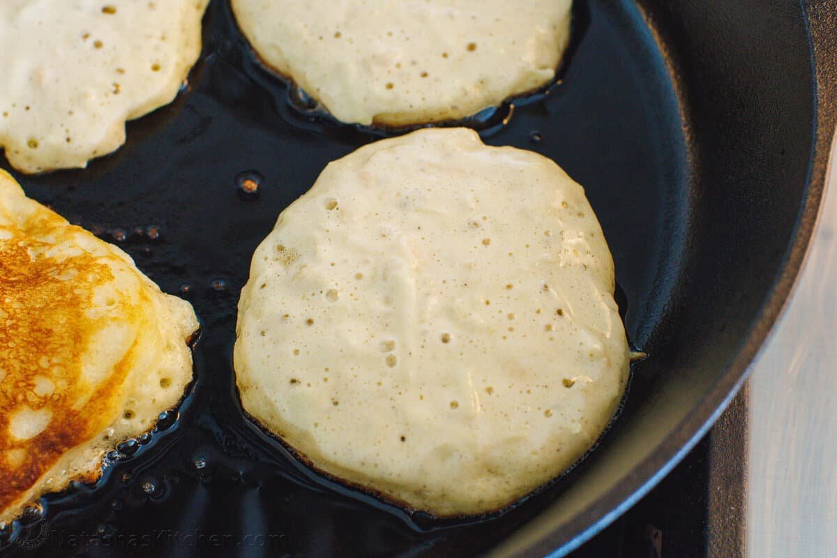 Apple pancakes cooking in a cast iron skillet, with bubbles showing that they're ready to be flipped.