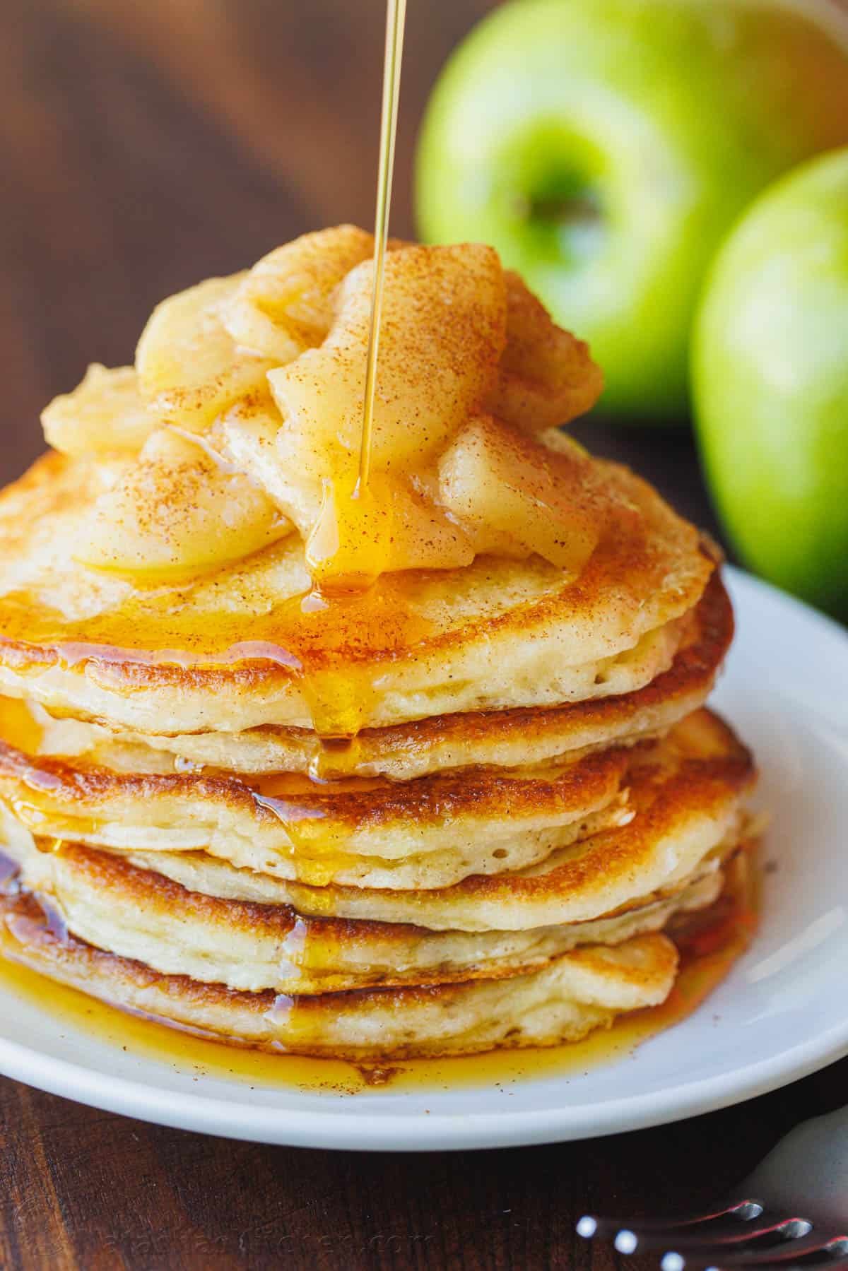 A stack of apple pancakes on a white plate, topped with sauteed cinnamon apples, with green apples in the background.