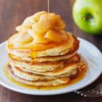 A stack of apple pancakes on a white plate, topped with sauteed cinnamon apples, next to a green apple.