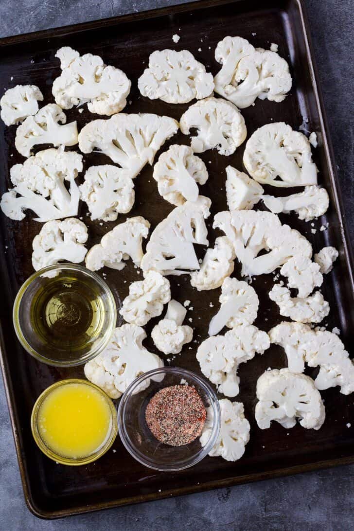 How to roast cauliflower in the oven with oil and seasonings. Cauliflower florets on a baking sheet with a glass bowl of oil and another bowl of seasoning.